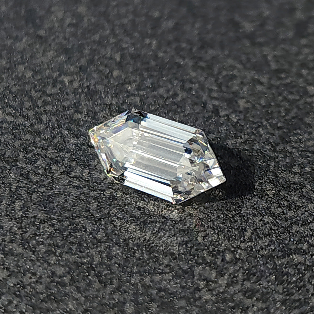 Elongated hexagon step-cut loose gem resting at an angle on a textured dark background showing light return.