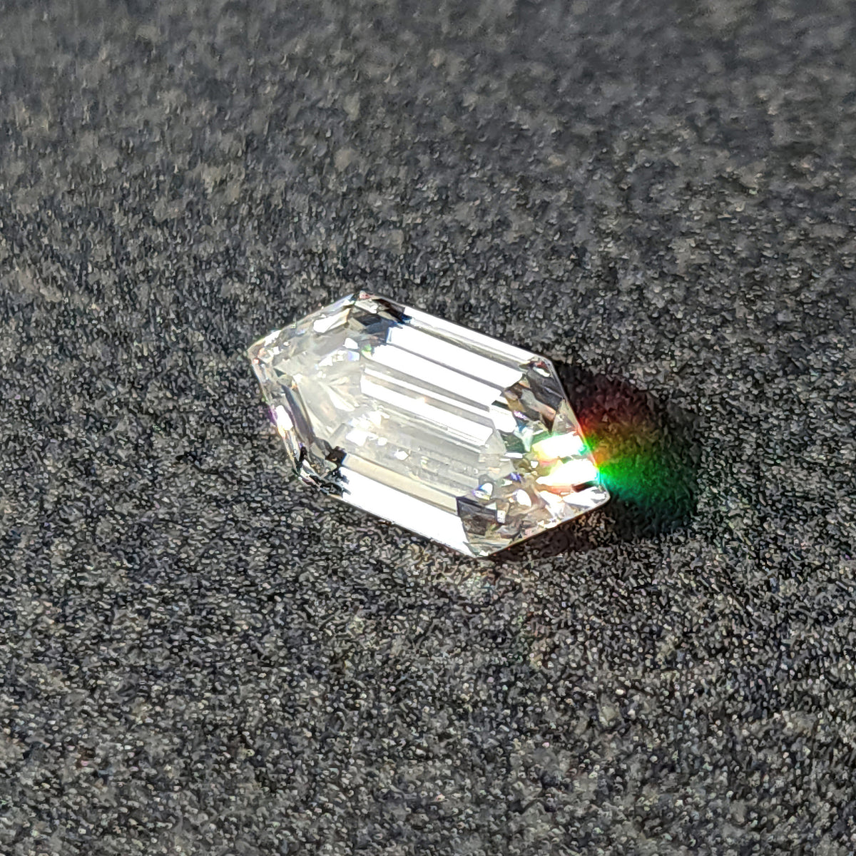 Elongated hexagon cut gemstone resting on a textured surface casting a distinct spectral dispersion patch.