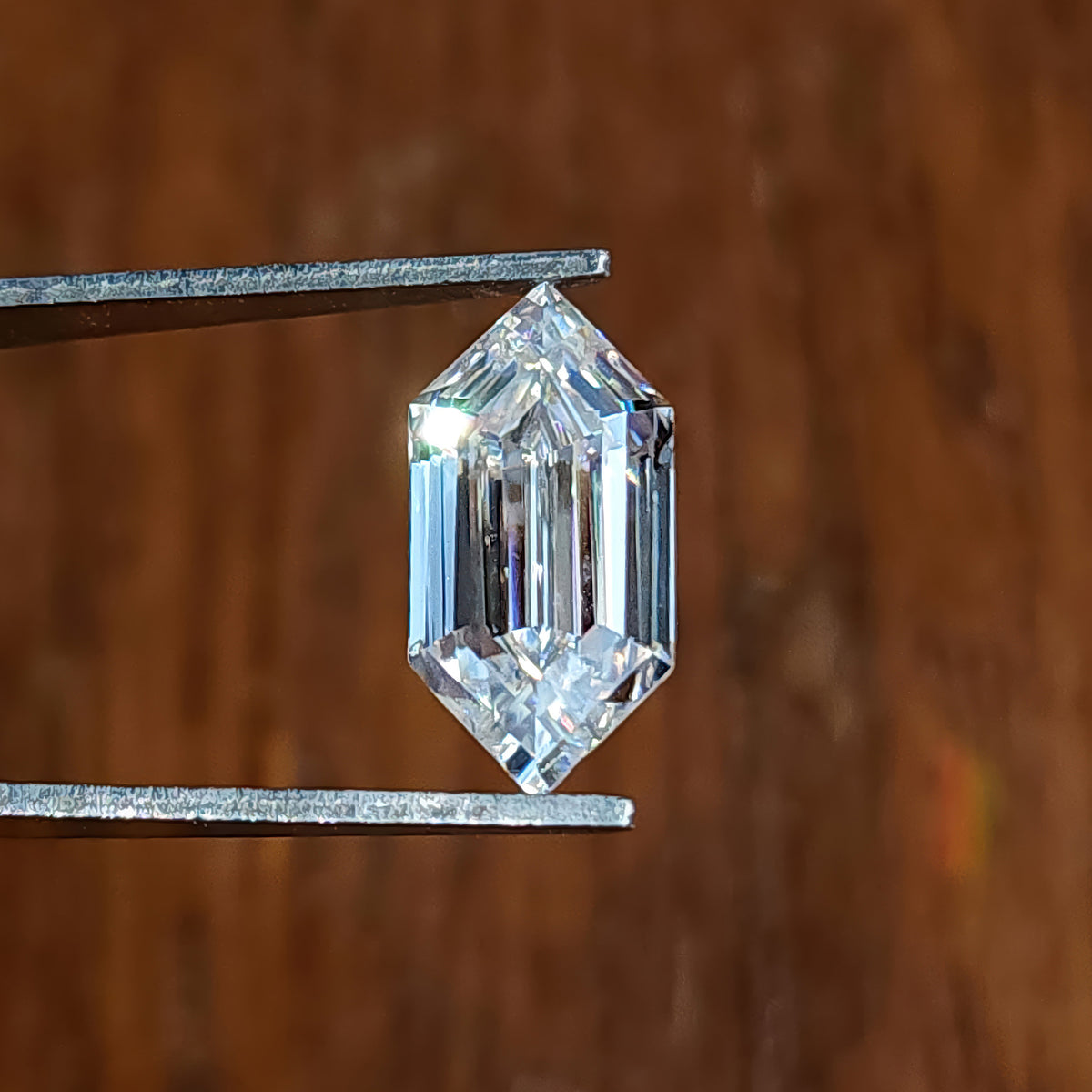 Colorless elongated hexagon step-cut gem held in tweezers over a warm vertical wood grain background.