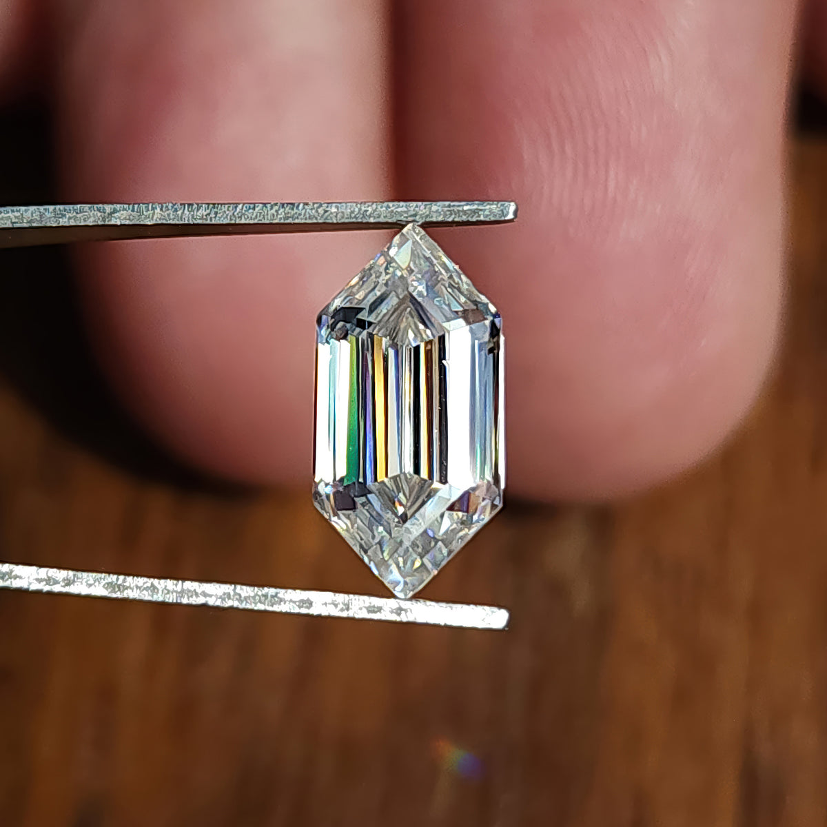 Elongated hexagon step-cut gemstone held in tweezers displaying colorful refraction bands.