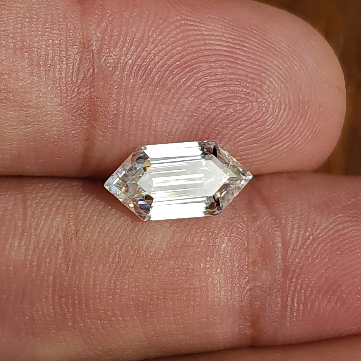Colorless elongated hexagon step-cut stone resting on skin under strong light showing table flash.