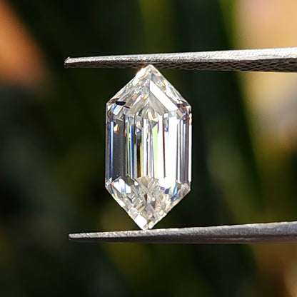Elongated hexagon step-cut gemstone held vertically in tweezers with diffuse green foliage bokeh behind.