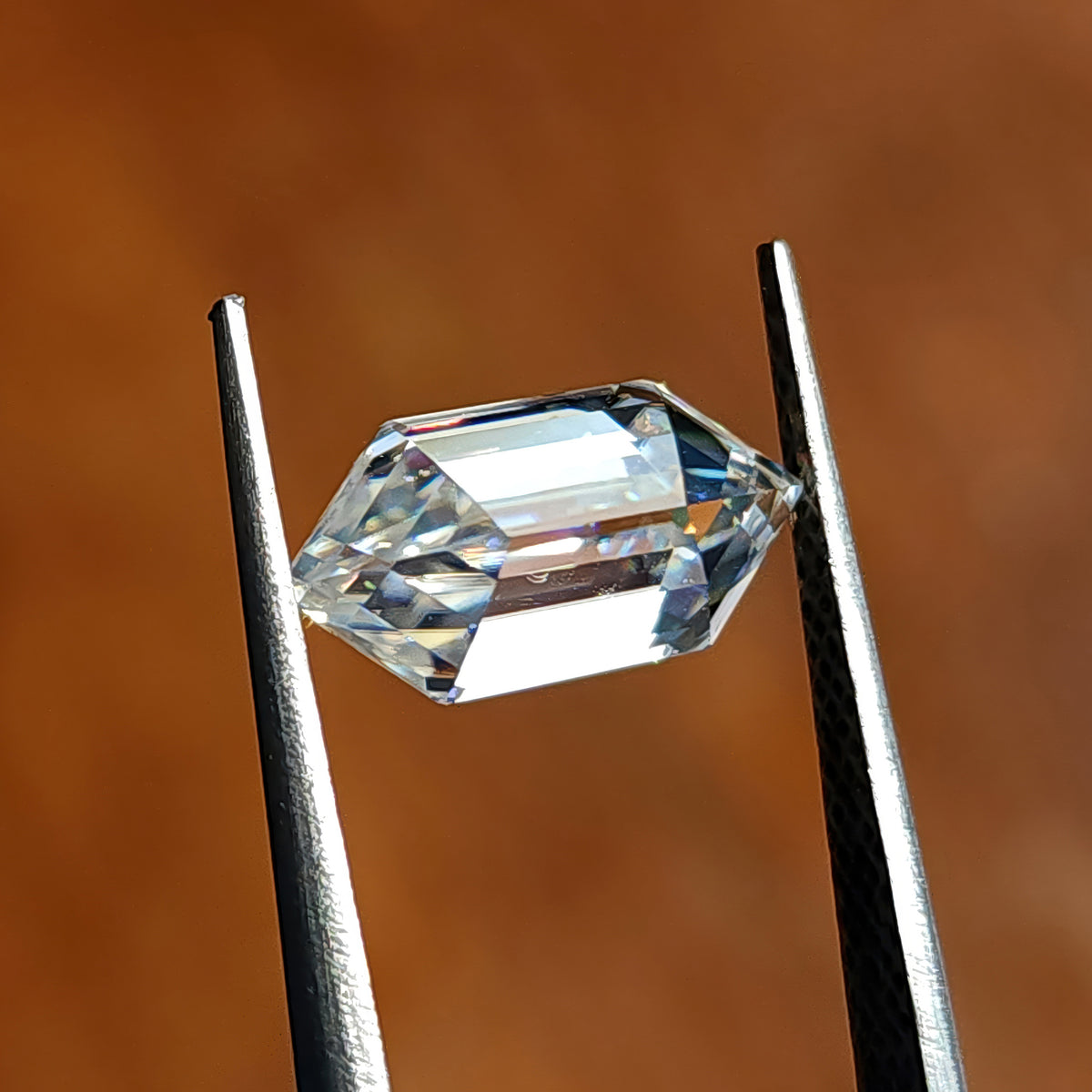 Elongated hexagon step-cut gem held horizontally across points in tweezers over warm brown ground.
