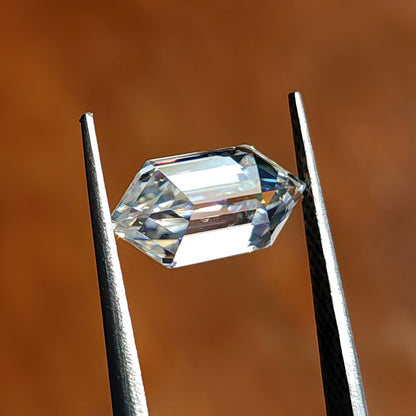 Elongated hexagon step-cut gem held horizontally across points in tweezers over warm brown ground.