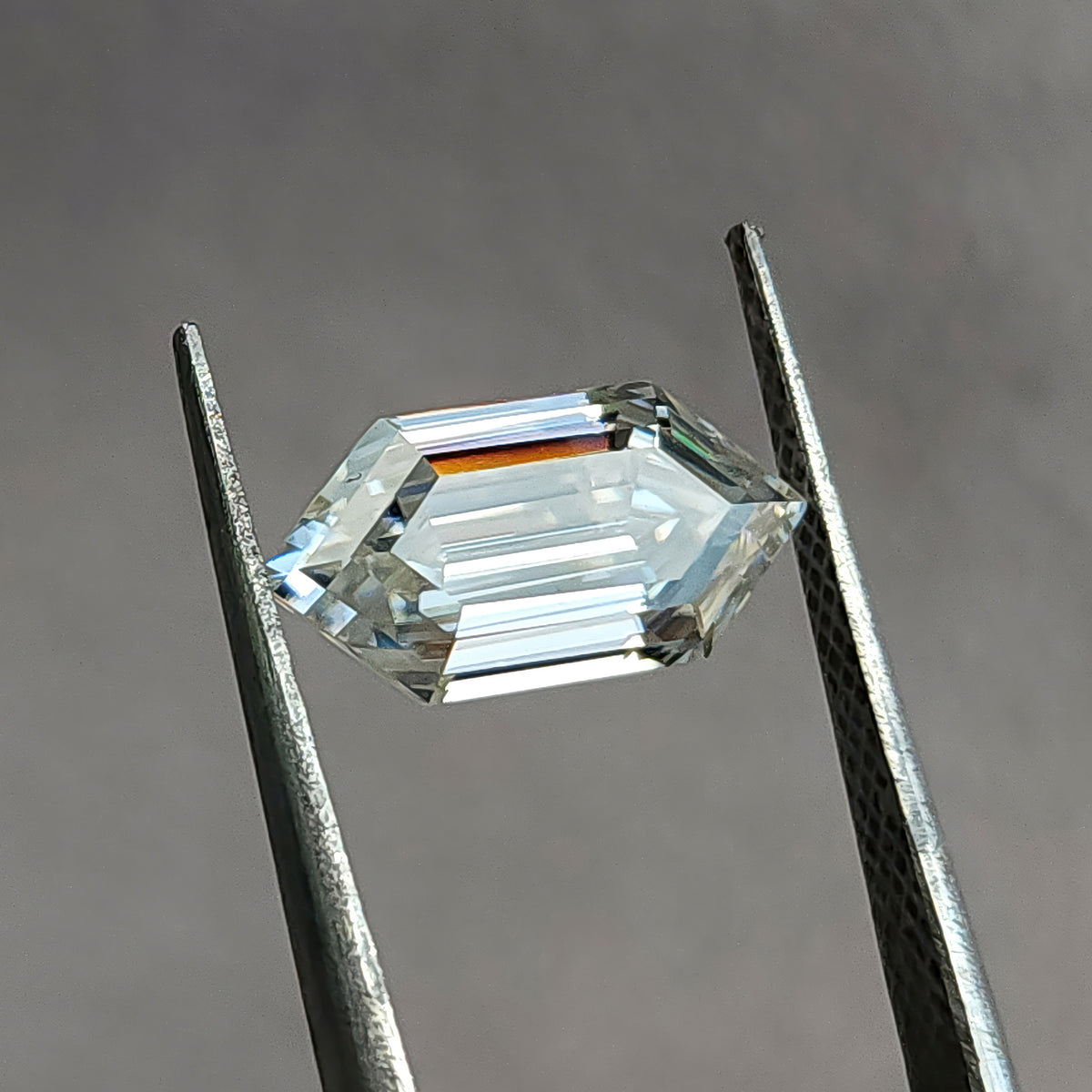 Colorless elongated hexagon step-cut gem held horizontally in tweezers showing symmetry.