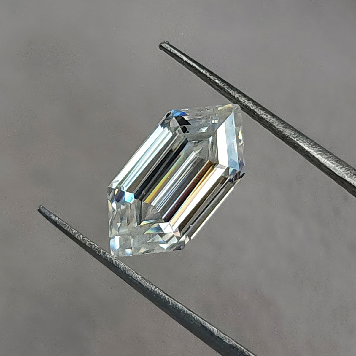 Elongated hexagon gemstone held diagonally in tweezers against a neutral grey background.