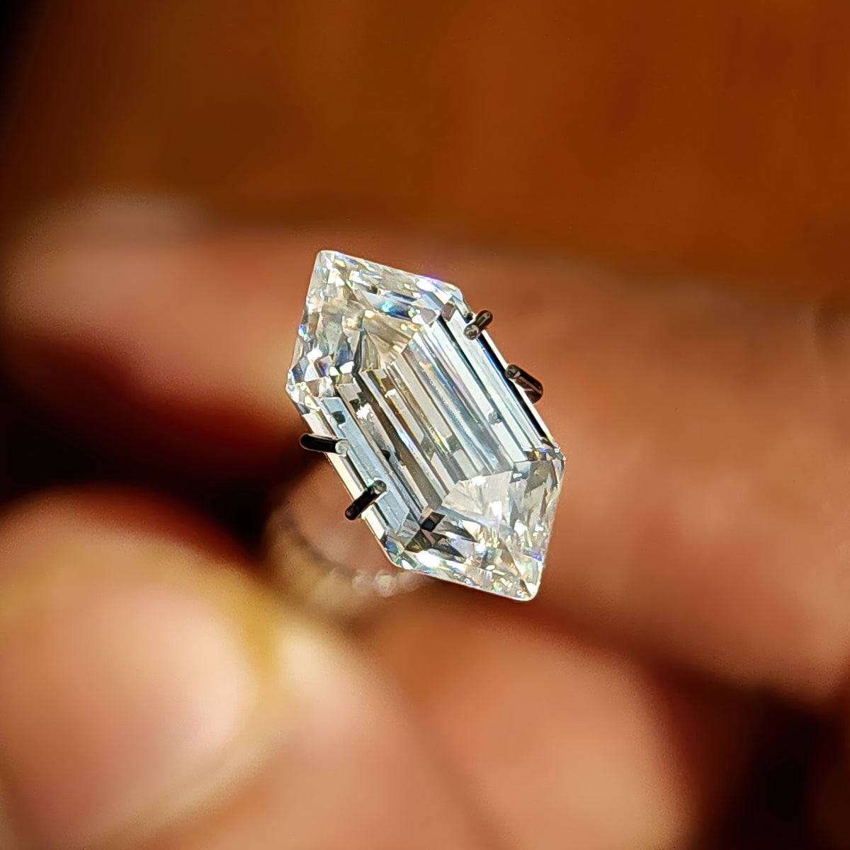 Elongated hexagon step-cut gemstone displayed in a claw prong setting holder on a hand.