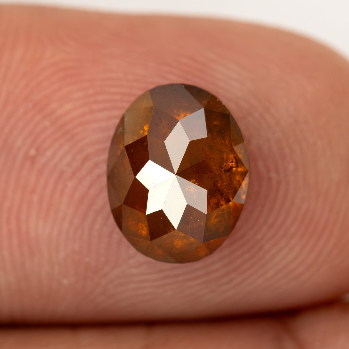 Oval reddish-orange diamond resting face-up on a fingertip for relative scale.