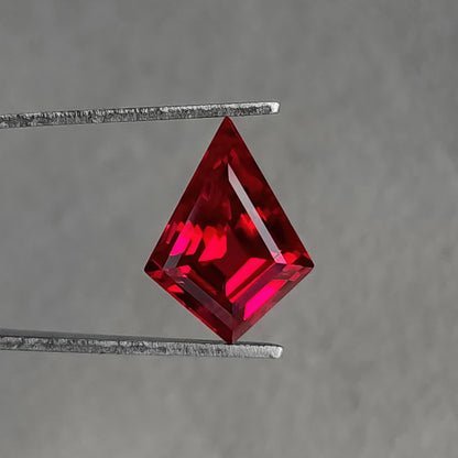 Face-up kite-shaped lab-grown ruby in tweezers against gray, with red and pink reflections and a bright diagonal right facet.
