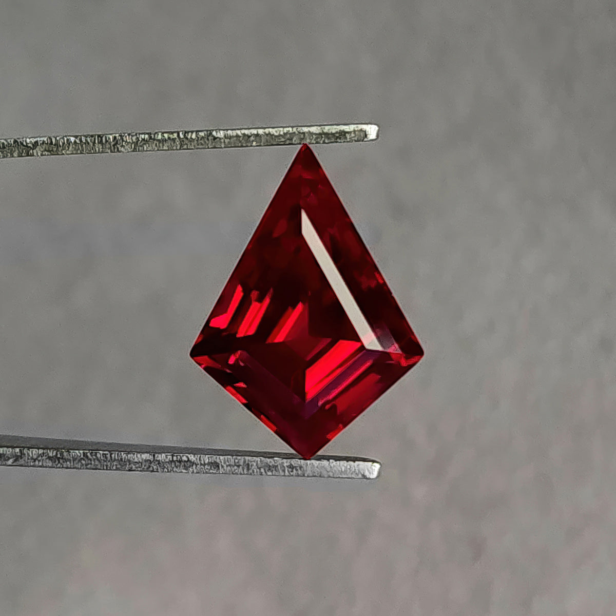 Face-up kite-shaped lab-grown ruby in tweezers, with bright red stripe-like reflections toward the left.