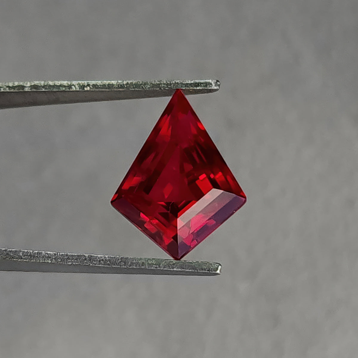 Tilted table-side kite-shaped lab-grown ruby in tweezers, with a dark central area and pale lower-right facet.