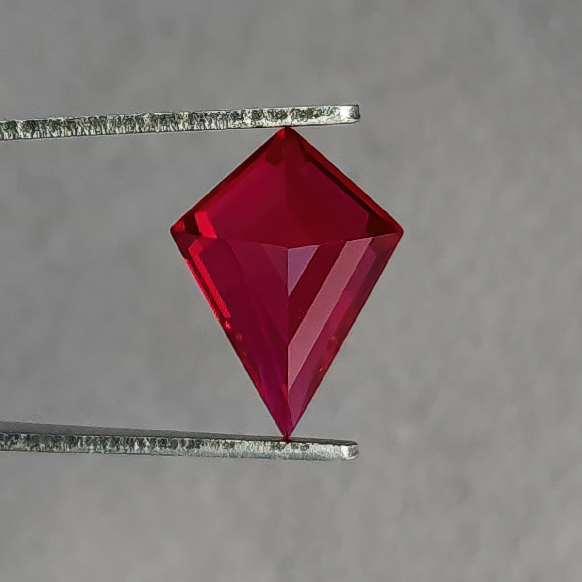 Reverse pavilion of a kite-shaped lab-grown ruby in tweezers, with the long point downward and dark red facets.