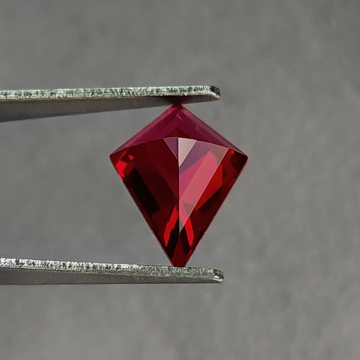 Reverse pavilion of a kite-shaped lab-grown ruby in tweezers, with the point downward, dark left facets and brighter right facets.