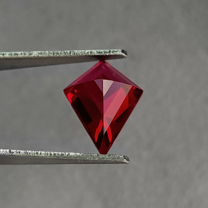 Reverse pavilion of a kite-shaped lab-grown ruby in tweezers, with the point downward, dark left facets and brighter right facets.