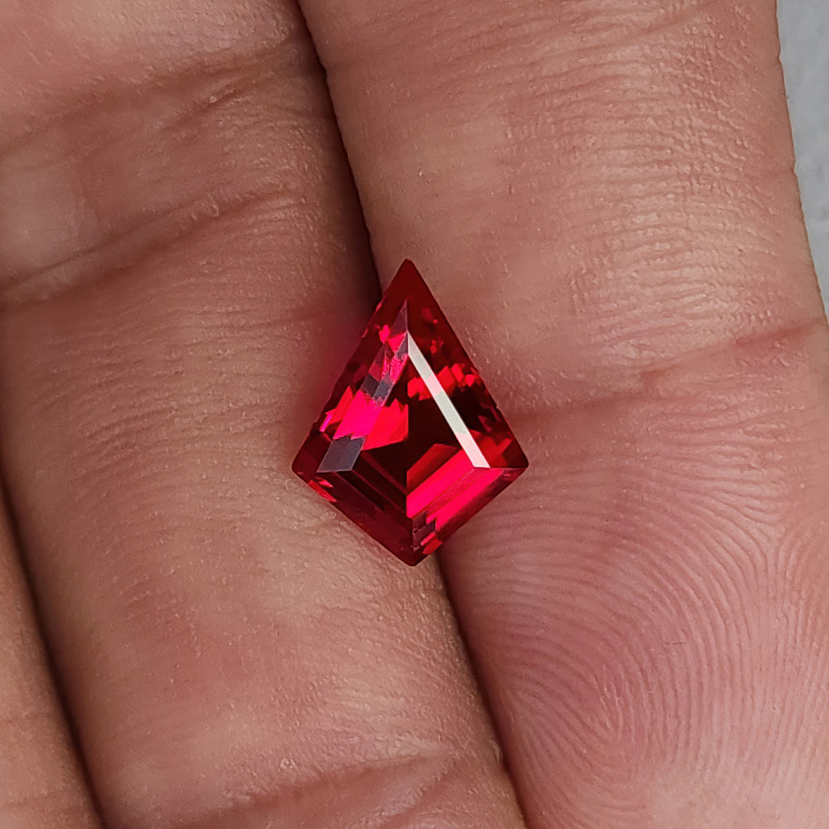 Face-up kite-shaped lab-grown ruby resting on fingers for scale, with red reflections and a thin bright right facet.