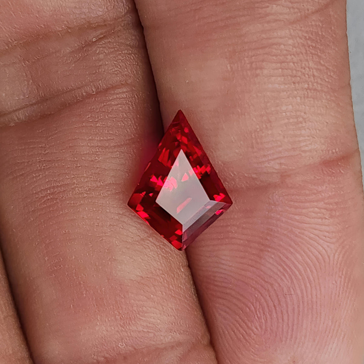 Kite-shaped lab-grown ruby on fingers for scale, with a broad pale pink reflection across the front table.