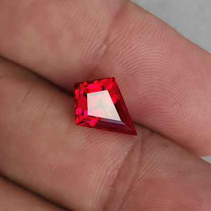 Rotated table-side kite-shaped lab-grown ruby on fingers, with the long point toward the lower right and a pale table reflection.