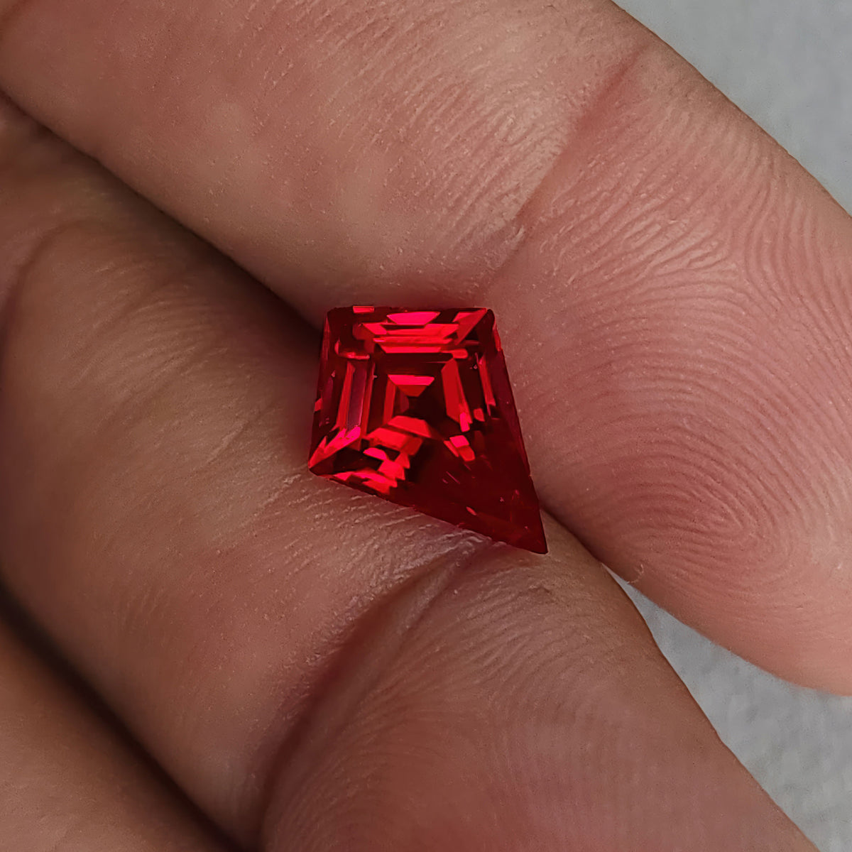Tilted kite-shaped lab-grown ruby on fingers, with a dark table and bright red nested facet reflections.