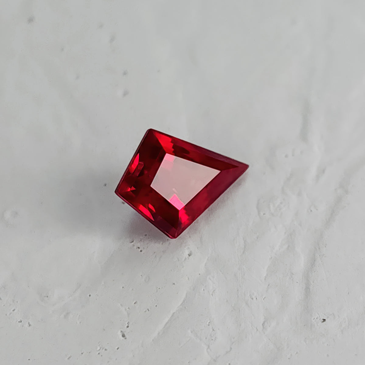 Table-side kite-shaped lab-grown ruby on a textured white surface, with the long point toward the lower right and a pale pink table.