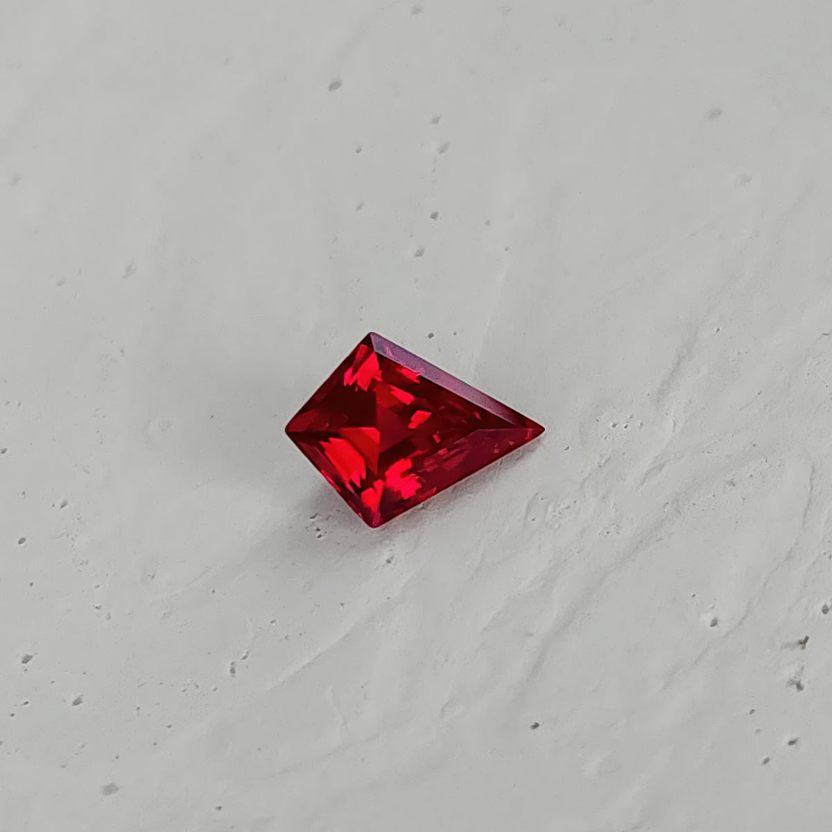 Kite-shaped lab-grown ruby on a textured white surface, showing darker front reflections and red facet borders.