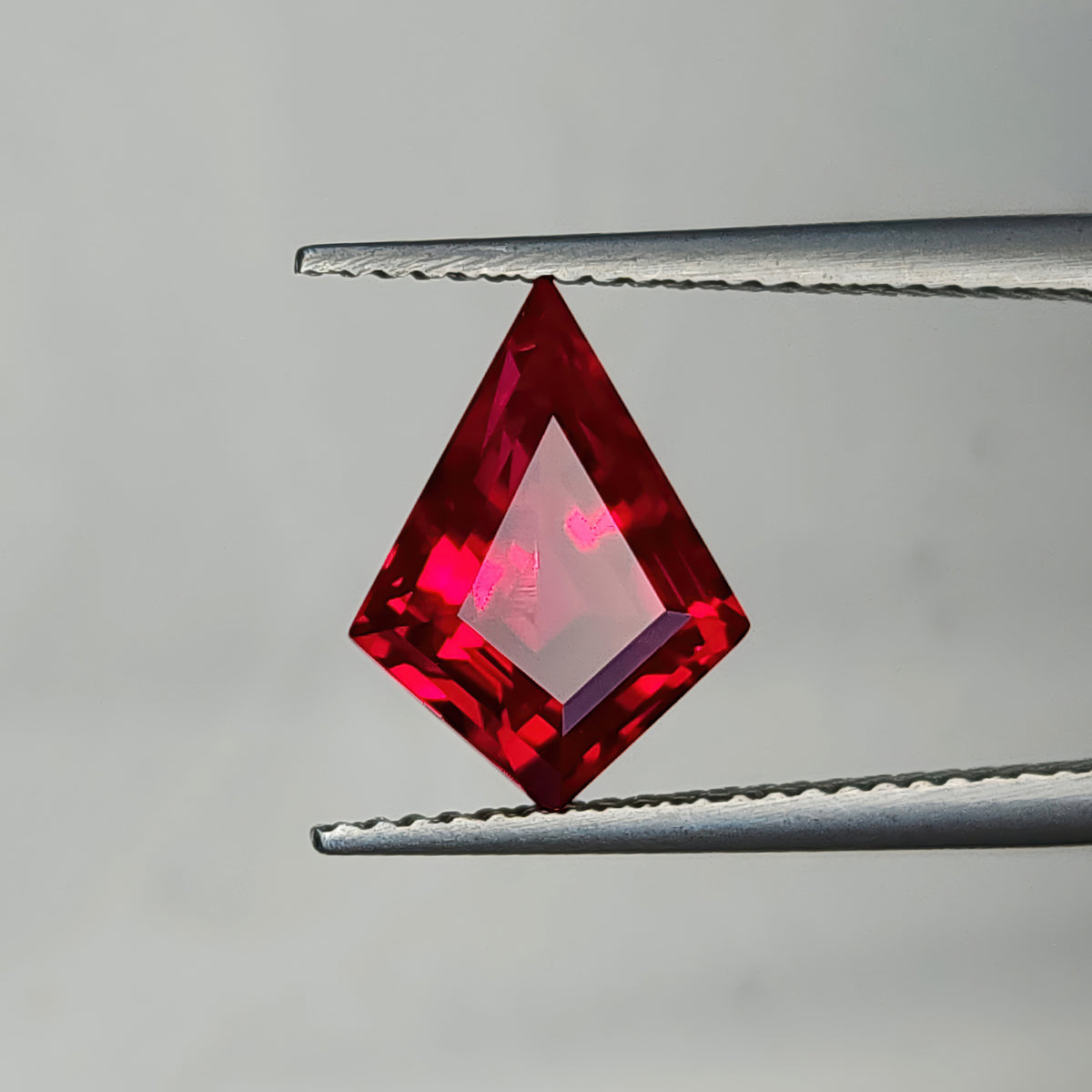 Face-up kite-shaped lab-grown ruby in tweezers against a light background, showing a large pale pink table reflection.
