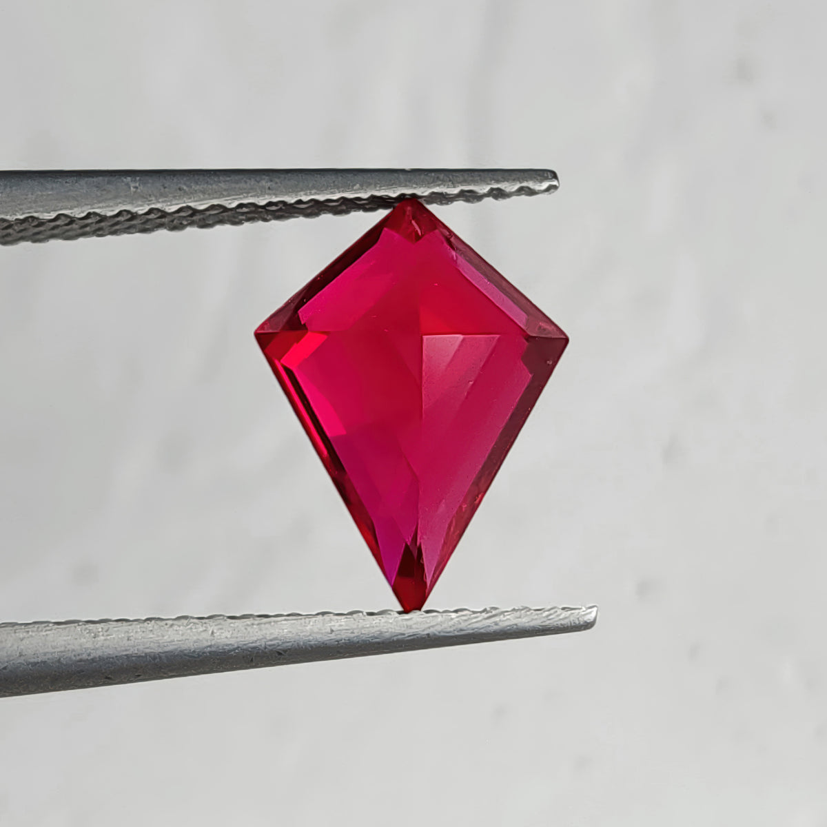 Reverse pavilion of a kite-shaped lab-grown ruby in tweezers against light gray, with bright pink facets along a central ridge.