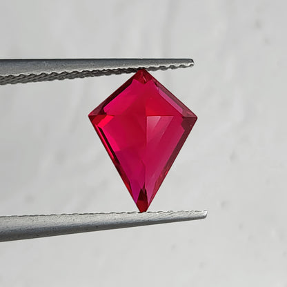 Reverse pavilion of a kite-shaped lab-grown ruby in tweezers against light gray, with bright pink facets along a central ridge.