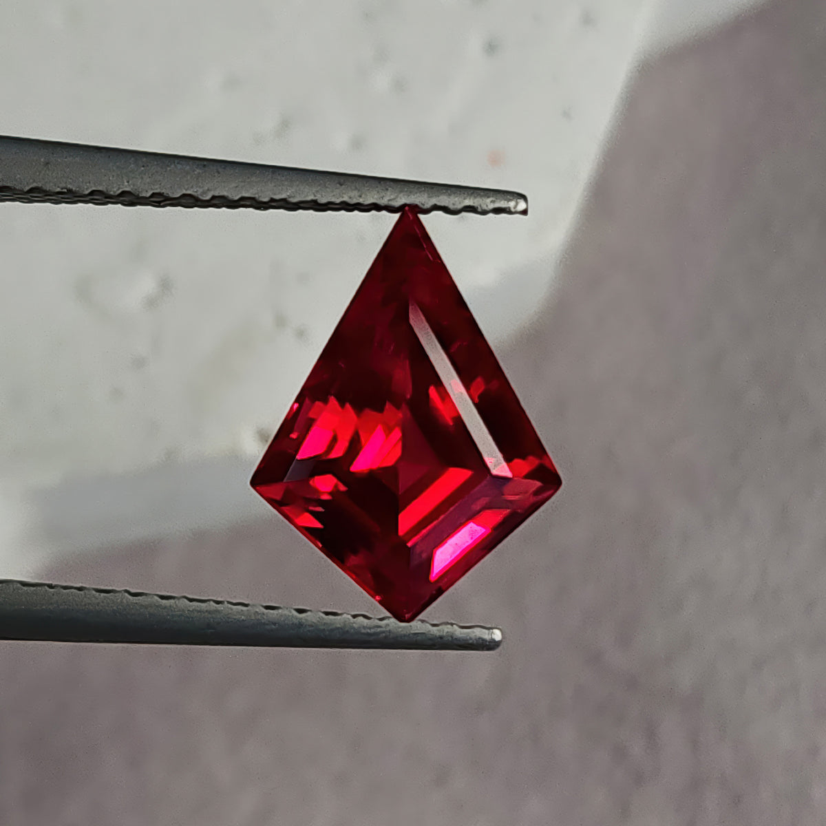 Face-up kite-shaped lab-grown ruby in tweezers against pale gray, with red-pink reflections and a bright diagonal right facet.