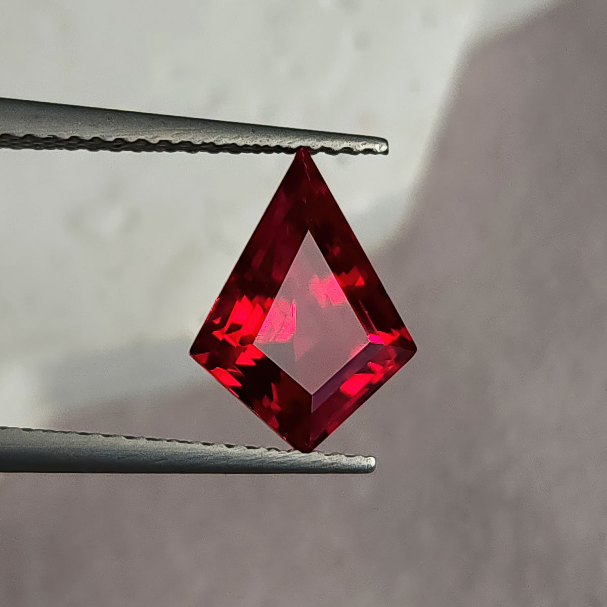 Table-side kite-shaped lab-grown ruby in tweezers against light gray, showing a broad pale pink flat table.