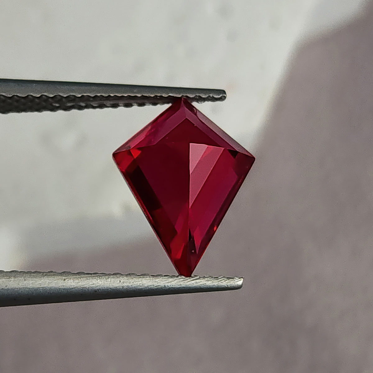 Reverse pavilion of a kite-shaped lab-grown ruby in tweezers against light gray, with dark left facets and bright red right facets.