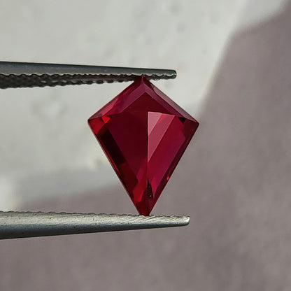 Reverse pavilion of a kite-shaped lab-grown ruby in tweezers against light gray, with dark left facets and bright red right facets.