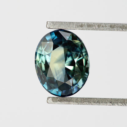 Oval blue-green sapphire held upright in tweezers, showing dark edge facets and a broad pale central reflection.
