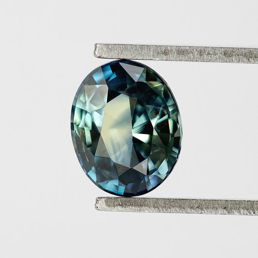 Oval blue-green sapphire held upright in tweezers, showing dark edge facets and a broad pale central reflection.