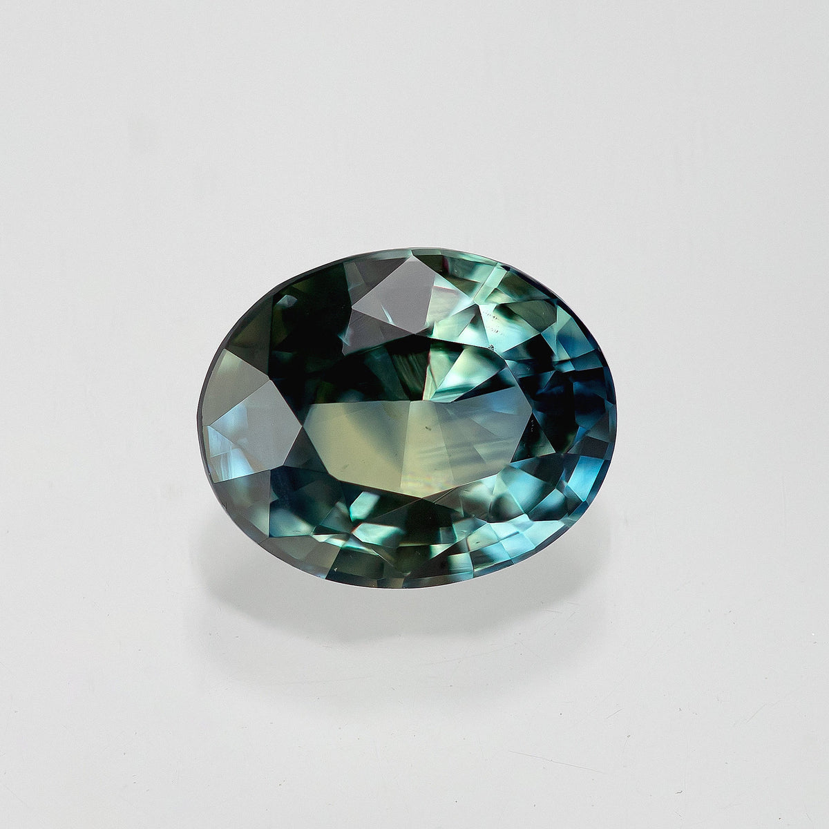 Oval blue-green sapphire lying horizontally on white, with dark upper facets and bright blue reflections at the right.