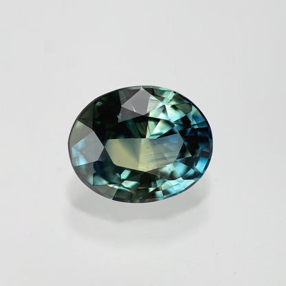 Oval blue-green sapphire lying horizontally on white, with dark upper facets and bright blue reflections at the right.