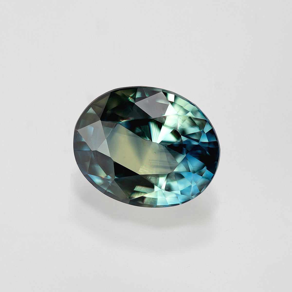Oval sapphire angled on a pale surface, showing blue-green reflections around a broad light central facet.