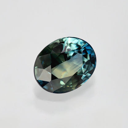 Blue-green oval sapphire tilted diagonally on white, showing dark facets at the upper edge and bright blue near the side.