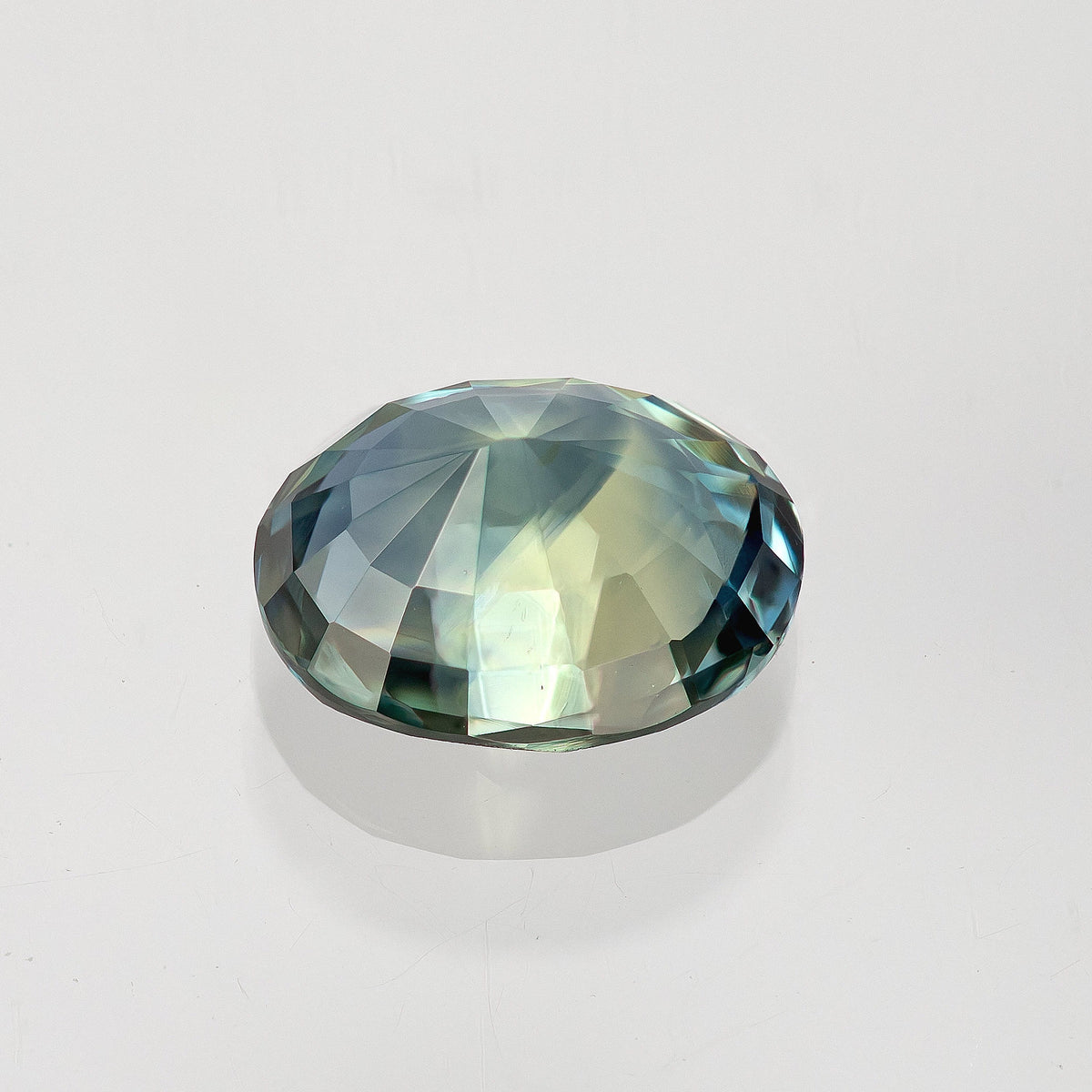 Pale blue-green pavilion view of the oval sapphire resting on white, showing the faceted back and curved outline.