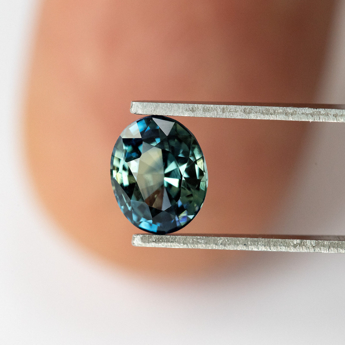 Oval blue-green sapphire held in tweezers in front of a fingertip, showing its size relative to the finger.