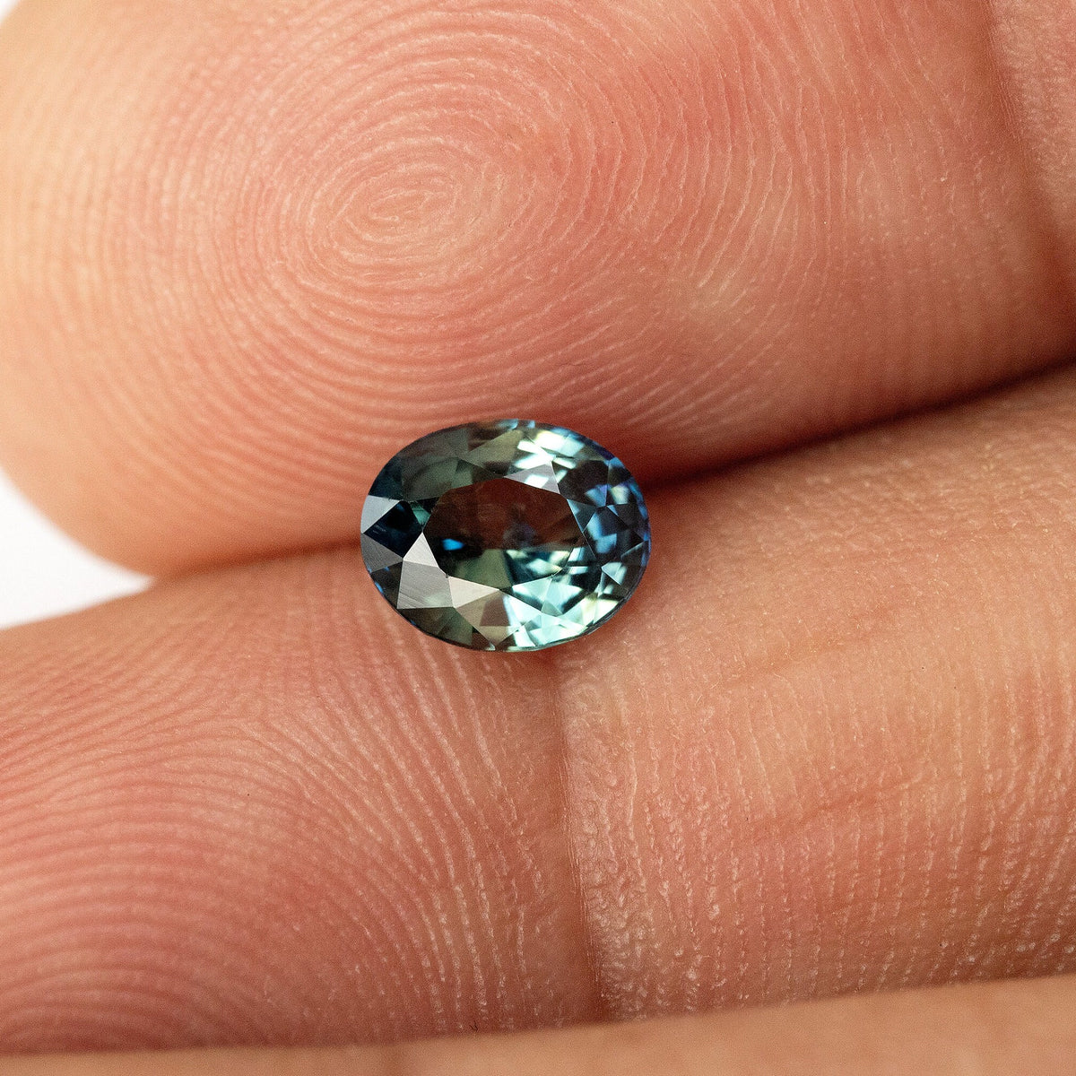 Small oval blue-green sapphire resting between fingertips, with the full outline and contrasting facet reflections visible.