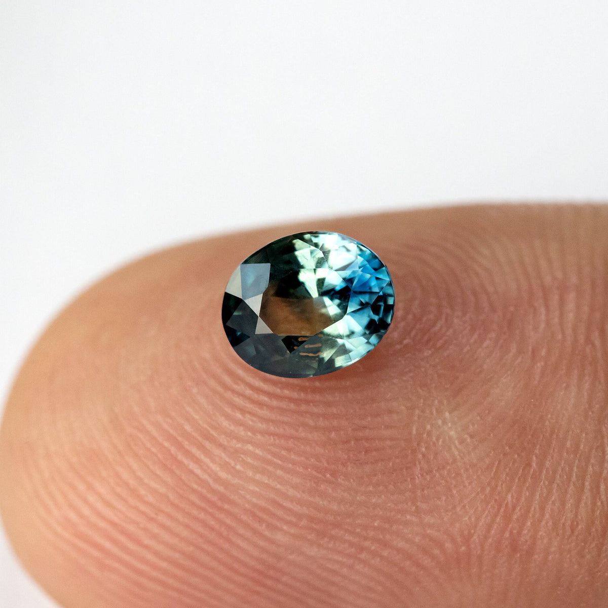 Oval sapphire resting face-up on a fingertip, with bright blue-green reflections across the upper crown.