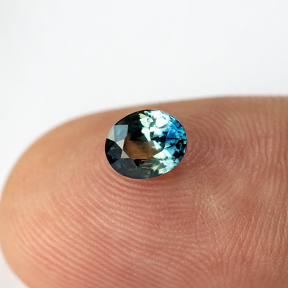 Oval sapphire resting face-up on a fingertip, with bright blue-green reflections across the upper crown.