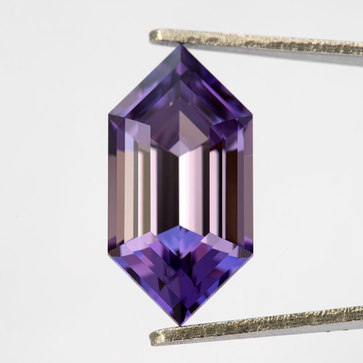 Zafiro hexagonal creado en laboratorio violeta púrpura visto desde arriba y sostenido verticalmente con pinzas contra blanco, con largas facetas escalonadas.