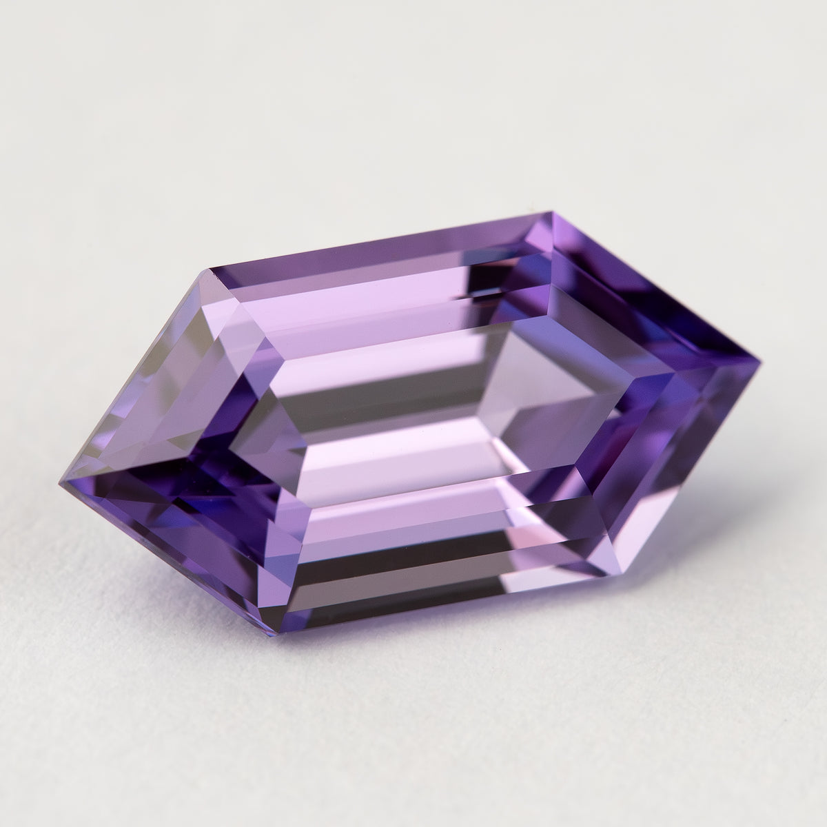Zafiro hexagonal violeta púrpura visto desde arriba en posición horizontal sobre blanco con bandas centrales pálidas y extremos puntiagudos más oscuros.