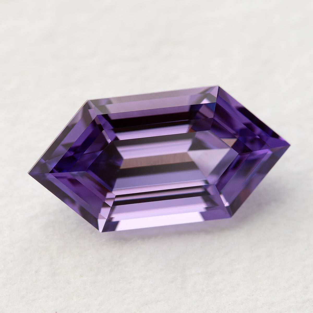 Zafiro hexagonal violeta púrpura visto desde arriba, descansando horizontalmente sobre blanco, mostrando bandas de facetas escalonadas alternas claras y púrpuras.