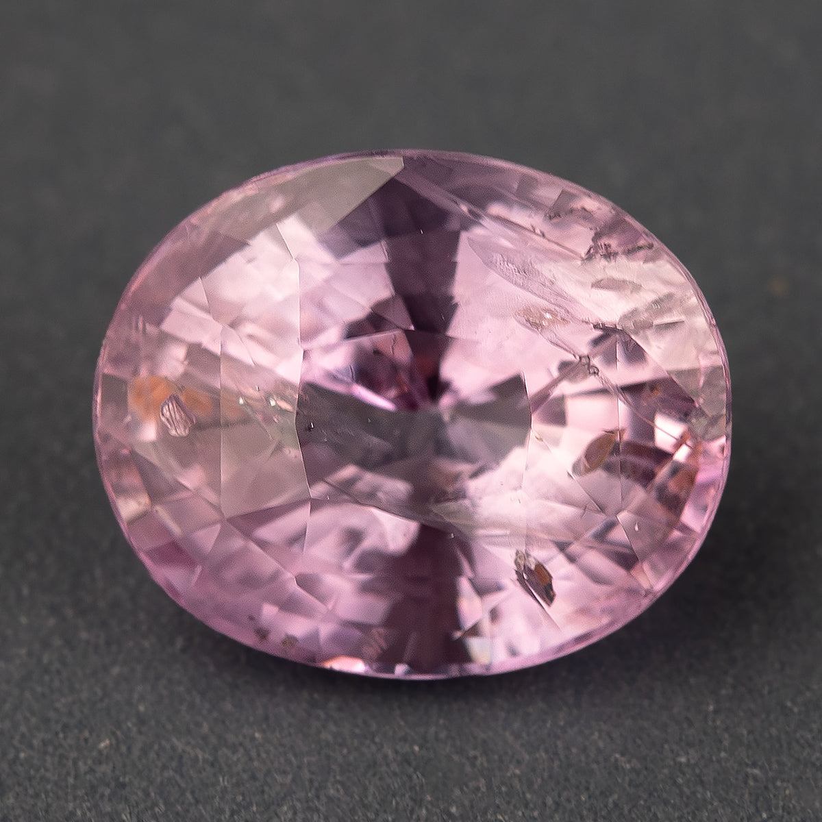 Pink oval sapphire face-up on a dark gray surface, showing its outline, facets and visible inclusions.