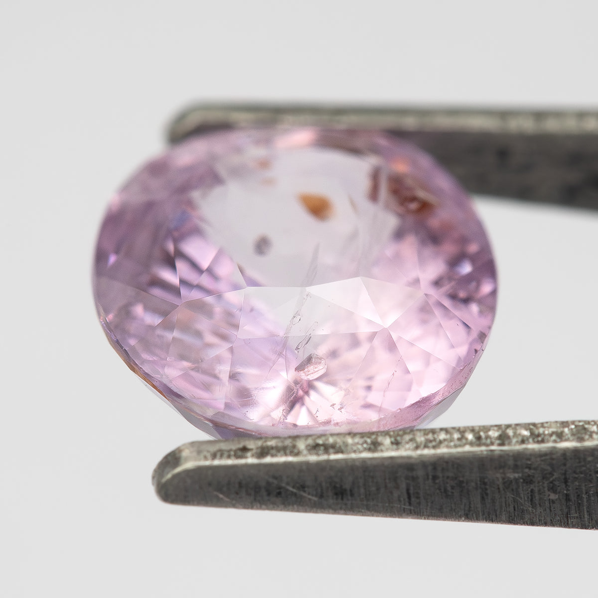 Reverse of the pink oval sapphire tilted in tweezers, showing pale reflections and orange-brown inclusions.