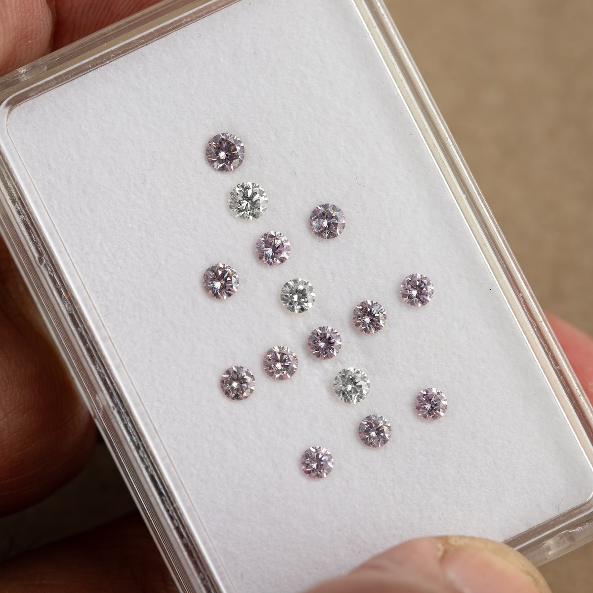 Close hand-held view of twelve purple-pink diamonds and three white comparison diamonds in a plastic box.