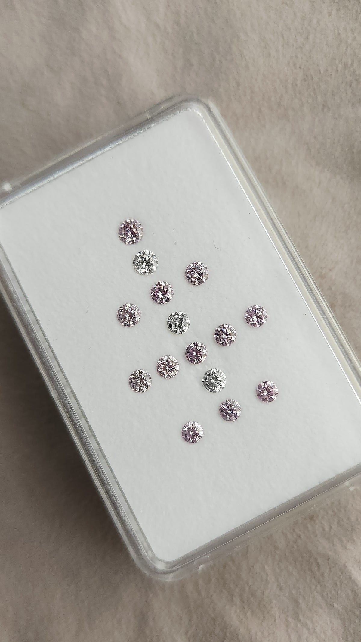 Angled view of twelve purple-pink diamonds and three white comparison diamonds in a plastic box.