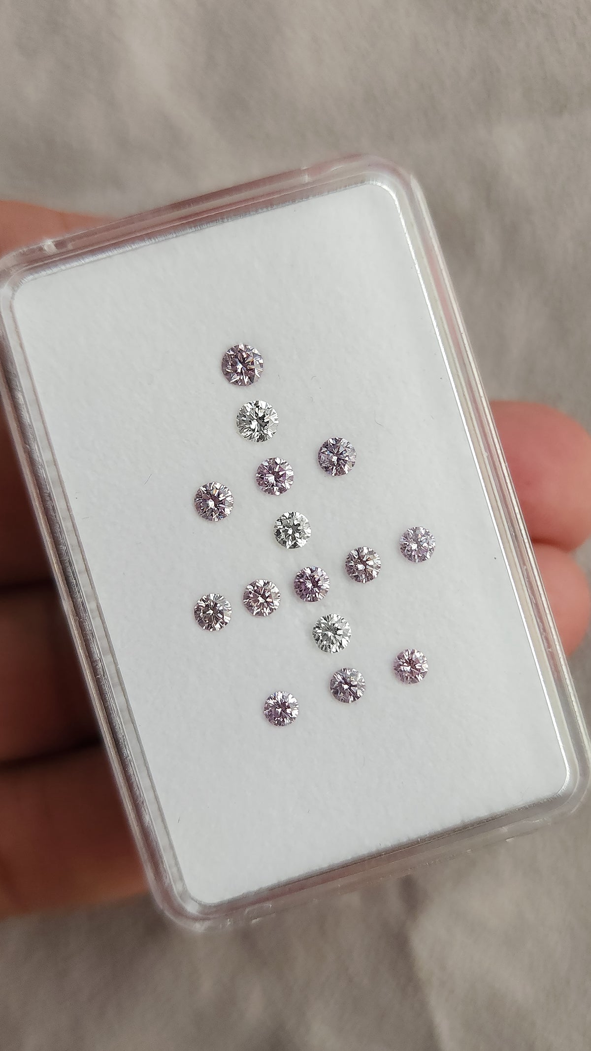 Hand-held view of twelve purple-pink diamonds and three white comparison diamonds in a plastic box.